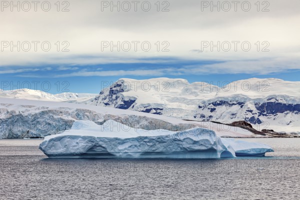 A large iceberg floats in the sea, surrounded by snowy mountains under a blue sky, iceberg in the