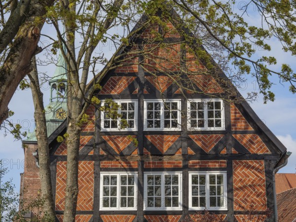 The picture shows a half-timbered house with red bricks, surrounded by trees. A church spire is