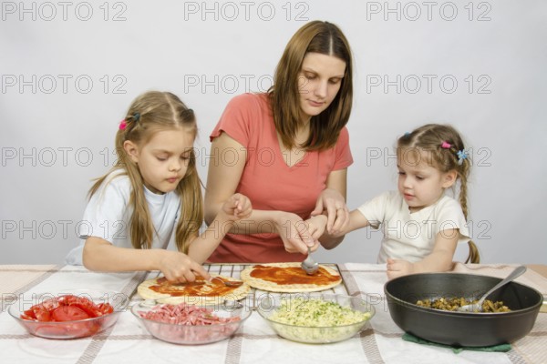 Mom helps younger daughter spread ketchup on a pizza, the eldest daughter, she is preparing a