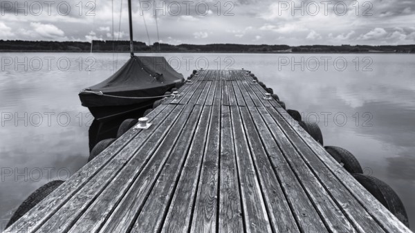 Wooden jetty and boat on a calm lake under a cloudy sky in black and white, Irsingener See, barrage