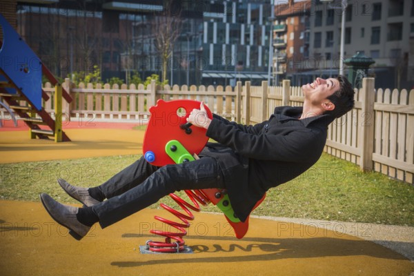 Young man reliving his childhood plying in a childrens playground riding on a colourful red spring