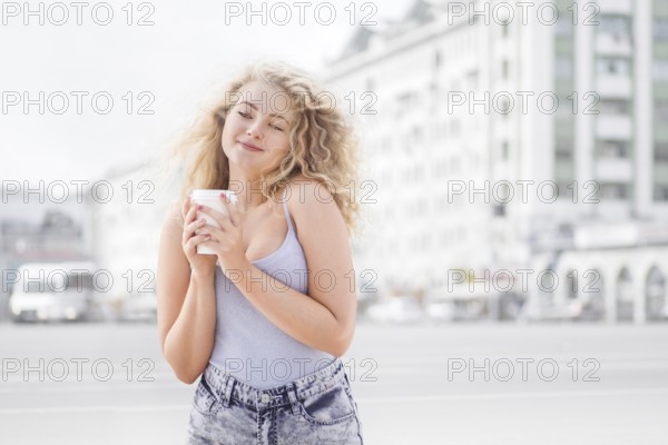 Happy young woman with long curly hair holding a takeaway coffee cup, smiling and flirting with the