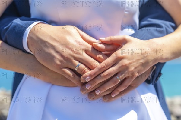 A couple is holding hands and wearing wedding rings. The woman is wearing a white dress and the man