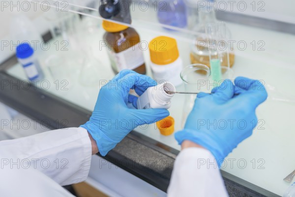 Person in a laboratory puts a small amount of powder into a beaker with a spatula, Youth Research