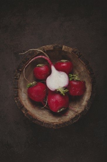 A collection of vibrant red radishes sits in a rustic wooden bowl. The contrasting dark background