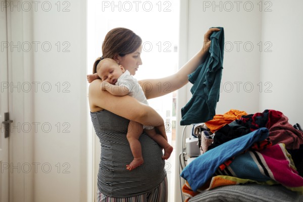 Young mother holding sleepeing baby on her shoulder and trying to sort the laundry simultaniously.