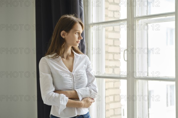 A teenager stands by a window, gazing outside with a thoughtful expression. The cozy room features