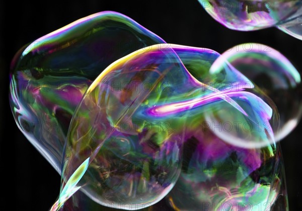 Large, irregular, iridescent soap bubbles in the colours of the rainbow against a black background,