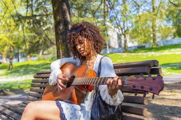Curly haired woman sitting on a bench playing an acoustic guitar in a green city park during a