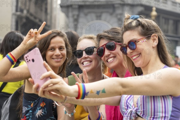 Four joyful women wearing colorful rainbow bracelets are capturing a selfie with a smartphone
