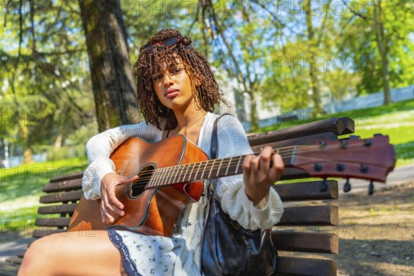 Young woman sitting on a park bench, playing an acoustic guitar while enjoying a sunny day filled