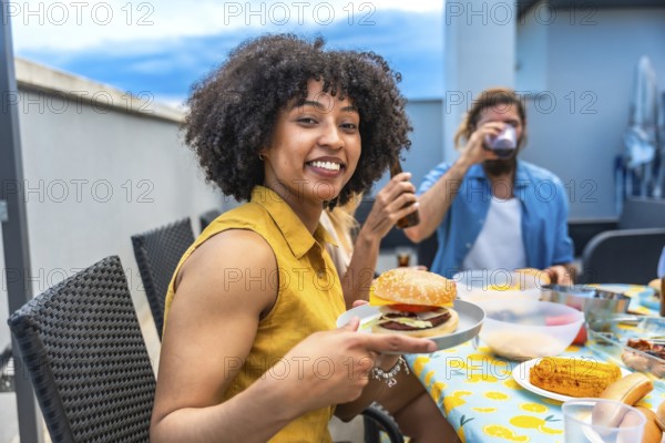 Young woman showing her delicious burger, enjoying a rooftop barbecue party with friends, drinking