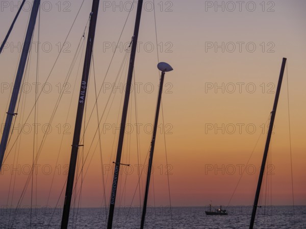 Masts in the foreground and a small boat in the sea at sunset, the sky is bathed in orange tones,