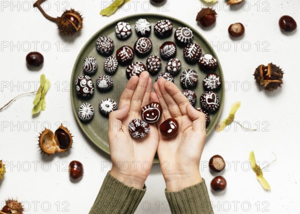 Two hands hold beautiful hand-painted chestnuts with different motifs over a plate full of autumn