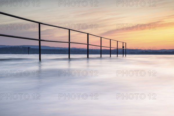 A railing in calm water, surrounded by a pastel-coloured sunrise, lido, Litzelstetten, Lake
