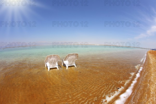 Two white beach chairs stood side by side in the clear water. The sun shines on the beach