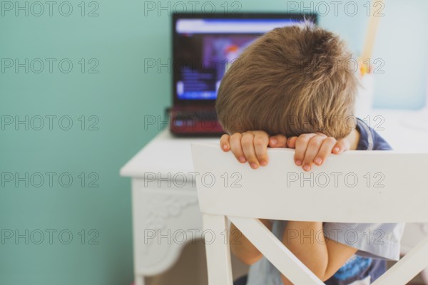 The sad little boy sits in front of laptop and doesn't want to look lesson. Concept of boring
