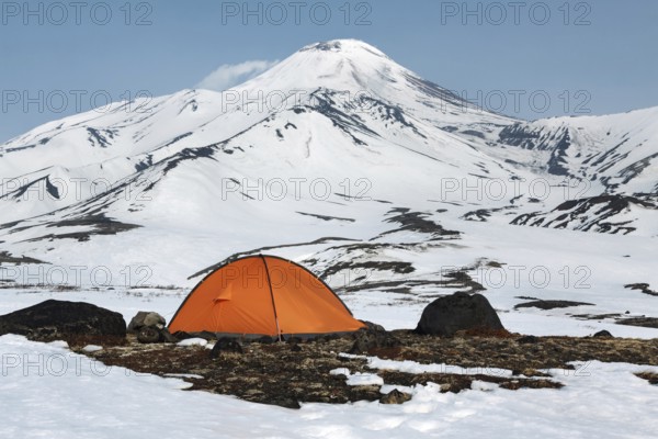 Orange camping (tourist) tent stands on meadow surrounded by snow at foot of beautiful active