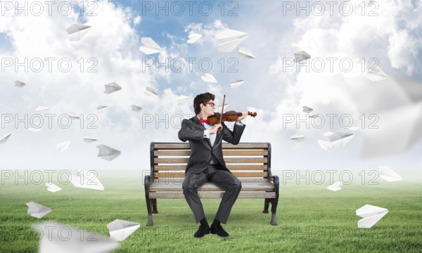 Young businessman plays the violin sitting on a bench, around flying paper planes. Creativity in