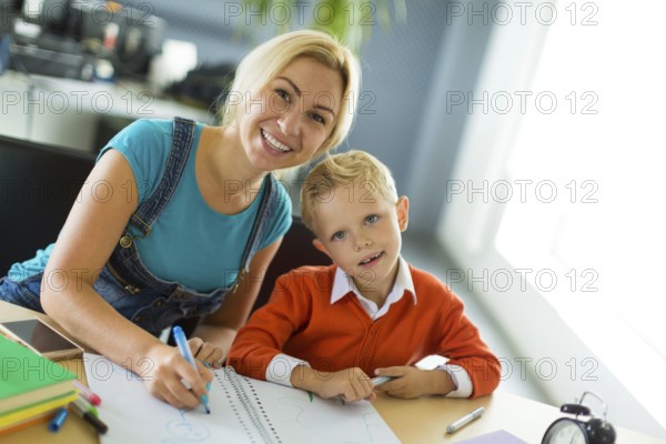 Young blonde caucasian kid in orange shirt and his mother sit at the table with colorful folders