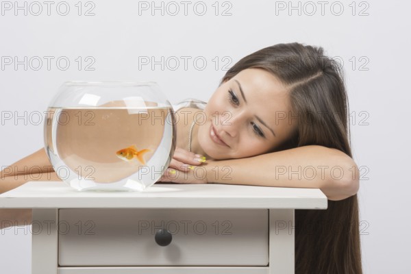 A young girl sits next to a round aquarium in which swimming goldfish