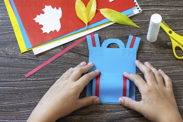 Instruction step 6. Greeting card paper backpack on a wooden table. Handmade. Project of children's