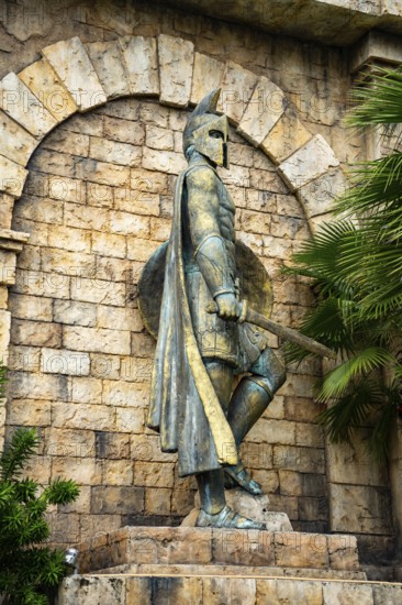 Close-up of an ancient statue with helmet and shield amidst stonework and greenery, palms, city,