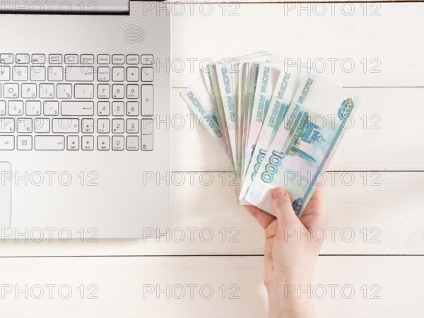 Hand holding russian rubles near laptop on white wooden background. Top view or flat lay. Finance