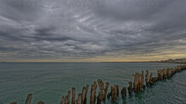 Calm sea with a row of wooden posts extending into the water under a dramatic cloudy sky,