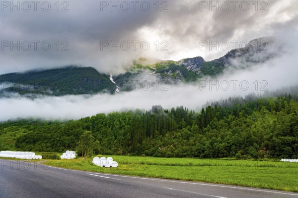 Wide asphalt road crosses the valley. Jostedalsbreen Park. Valley surrounded by high forested