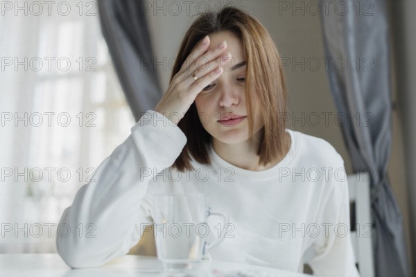 Woman holding her forehead looking fatigued, sitting indoors with a glass of water, suggesting a