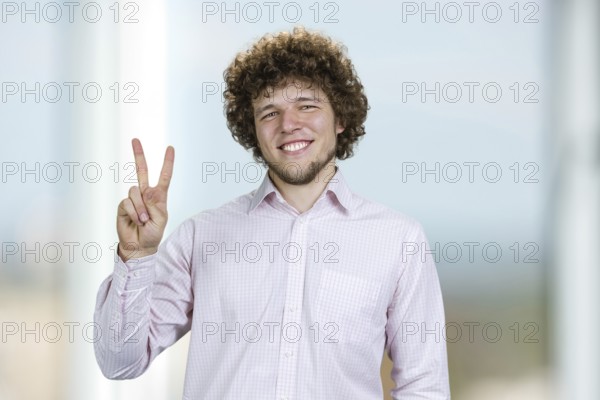 Portrait of a happy young man with curly hair shows victory gesture sign. Indoor window in the
