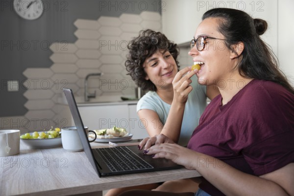 A happy lesbian couple enjoys feeding each other while working. Their laughter, casual attire, and