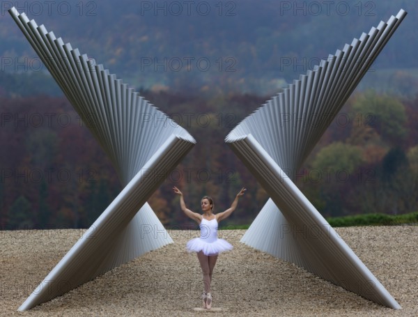 Ballerina in white ballet costume in elegant pose in front of modern sculpture in autumnal