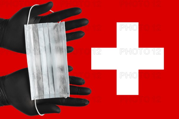 Doctor holds face mask in hands in black medical gloves on background colors flag of Switzerland.