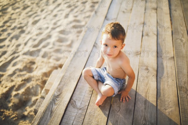Little Caucasian boy child sitting on wooden pier sandy beach, summer time, sea holiday near water.