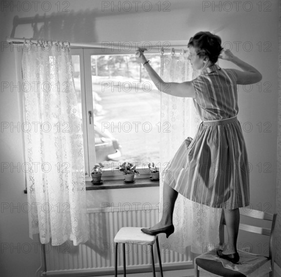 1960s home decorating. A woman at home putting up the curtains in the room window. All by herself