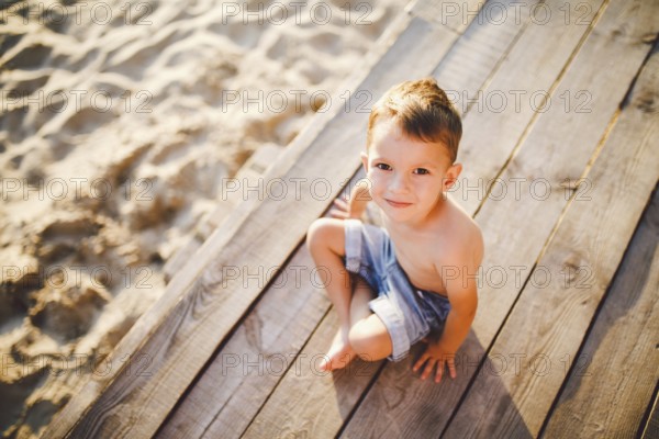 The theme is a child and summer beach holiday. A small Caucasian boy sits sideways on a wooden pier