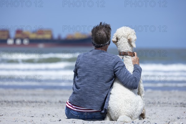 Woman sits together with her dog, a white poodle, amicably familiar on the beach in the sunshine,