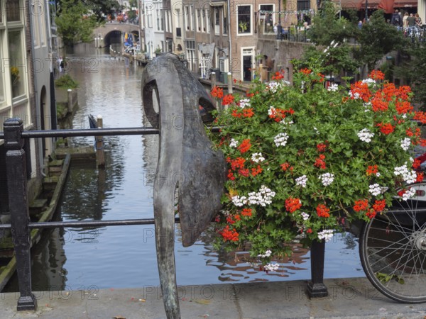 Sculpture and bicycle on a railing along a canal with blooming flowers and reflecting buildings,