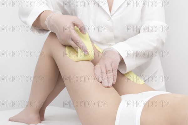 Unrecognizable beautician in white coat removal hair on women legs. Depilation procedure with green