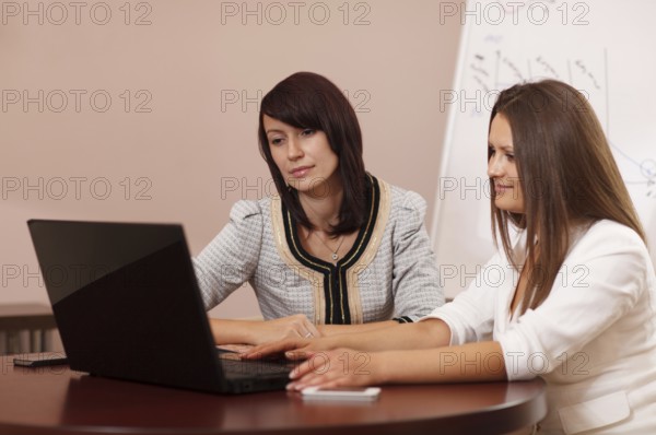 Two attractive stylish young women sitting at a table with a laptop computer reading something on