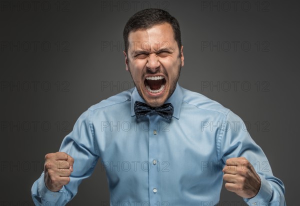 Portrait of angry upset young man in blue shirt and butterfly tie with fists up yelling isolated on