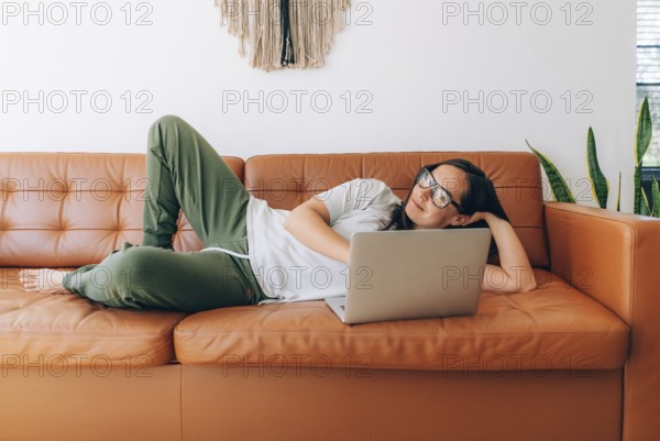 Junge glückliche Frau mit Brille, die zu Hause auf der Couch liegt und an ihrem Laptop arbeitet