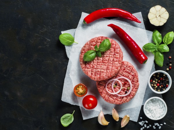 Two raw beef meat steak cutlets for burger with spices and vegetables on black concrete background.