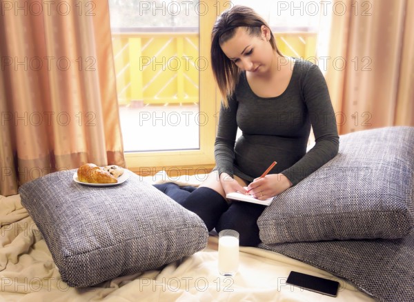 Beautiful pregnant woman sits comfortable on pillows, in front of big windows, with snacks nearby