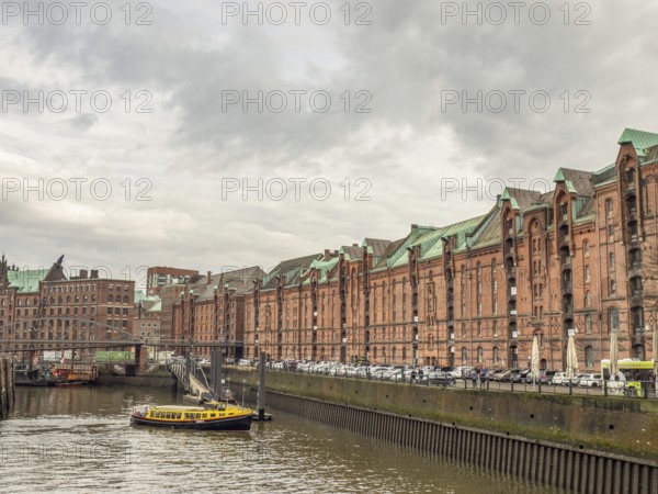 Yellow boat on a river next to historic brick-red buildings and a metal bridge under a cloudy sky,