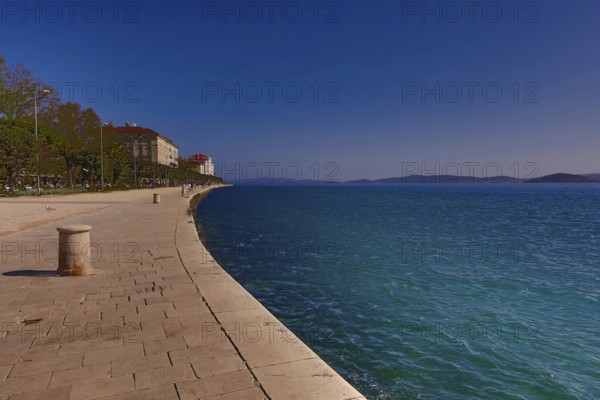 Long promenade along the blue water, with historic buildings and a clear sky in the background,