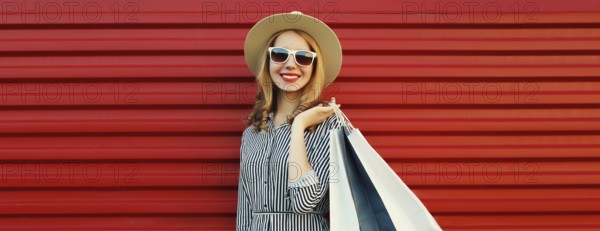 Stylish beautiful happy smiling young woman posing with shopping bags in summer straw hat on red