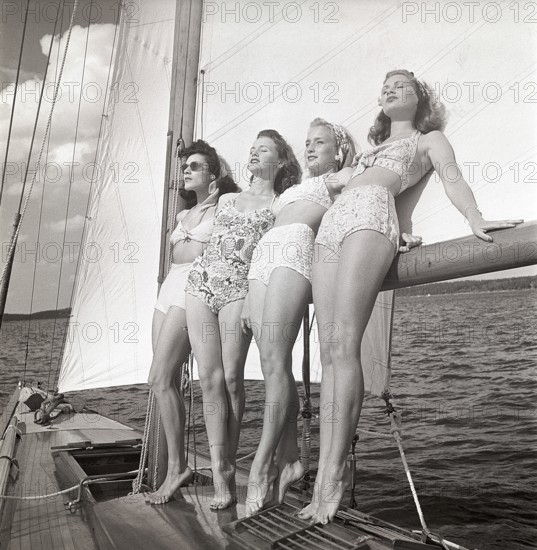 1940s sailing boat. Four young women are standing and leaning against the sails of the boat and